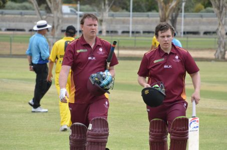 two male cricket players wearing maroon uniform and knee pads, carrying helmets and cricket bats in hand.