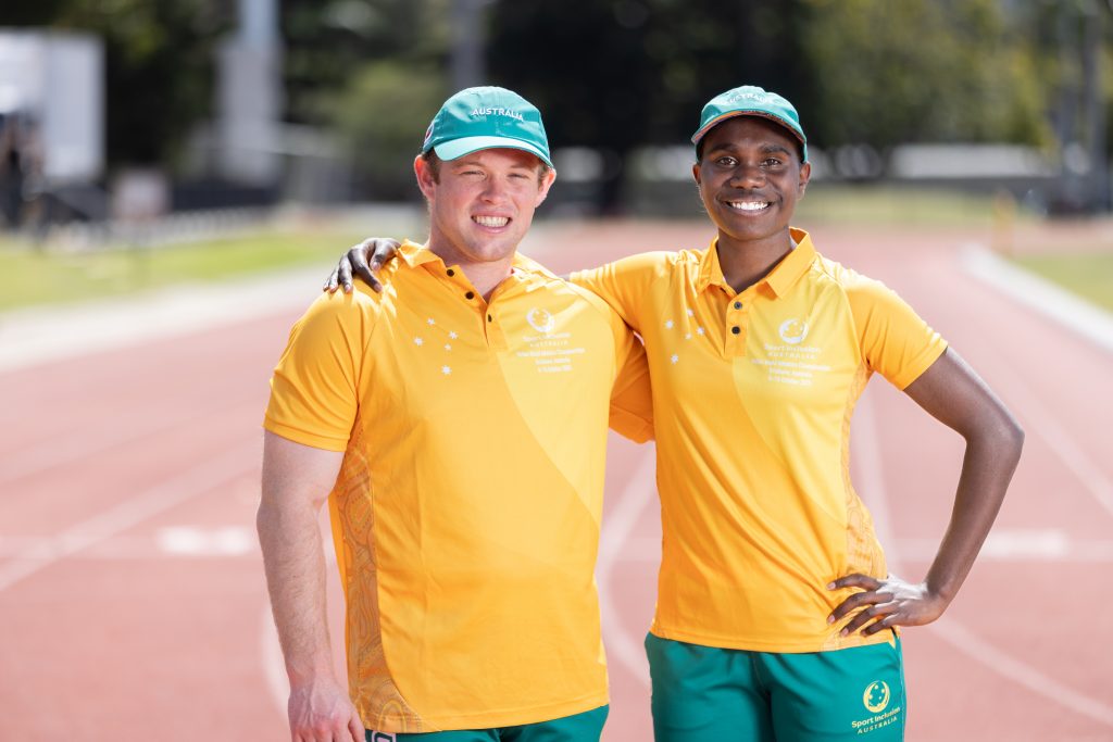 Two Australian athletes, Patrick Ramm (left) and Telaya Blacksmith (right) stand together smiling in their green and gold Australian Uniforms at an athletics track.